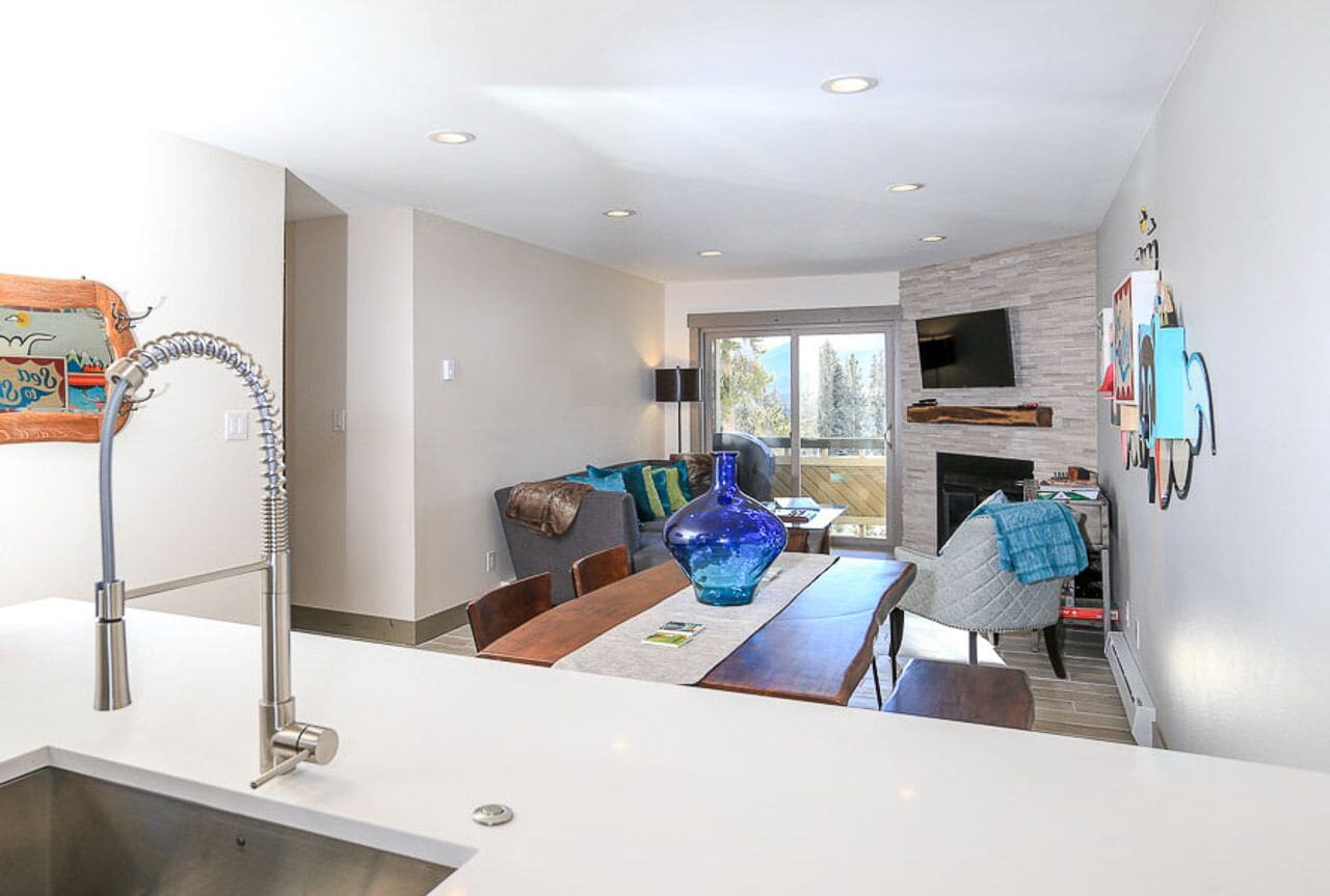 A modern kitchen with a stainless steel sink, a dining area with a wooden table and chairs, a blue vase, and a fireplace.