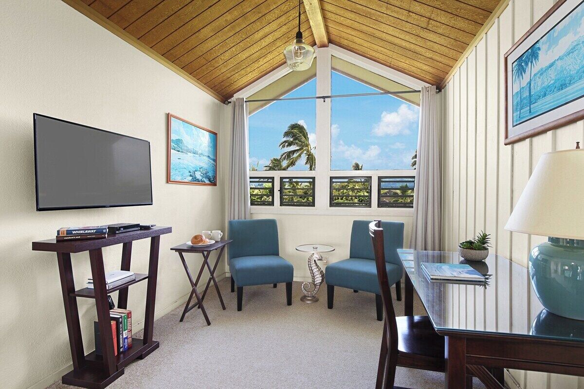 Een kamer met een televisie, twee blauwe stoelen, een kleine tafel, een lamp en een raam met uitzicht op palmbomen.