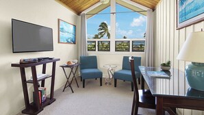 Una habitación con televisión, dos sillas azules, una mesita, una lámpara y una ventana que da a palmeras.