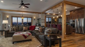 A spacious living room with a large sofa, ottoman, and a dining area with chairs. A kitchen with modern appliances is visible in the background. - Magnificent + Hot Tub + Close to Skiing - Falconhead Lodge - North (Steamboat Springs)