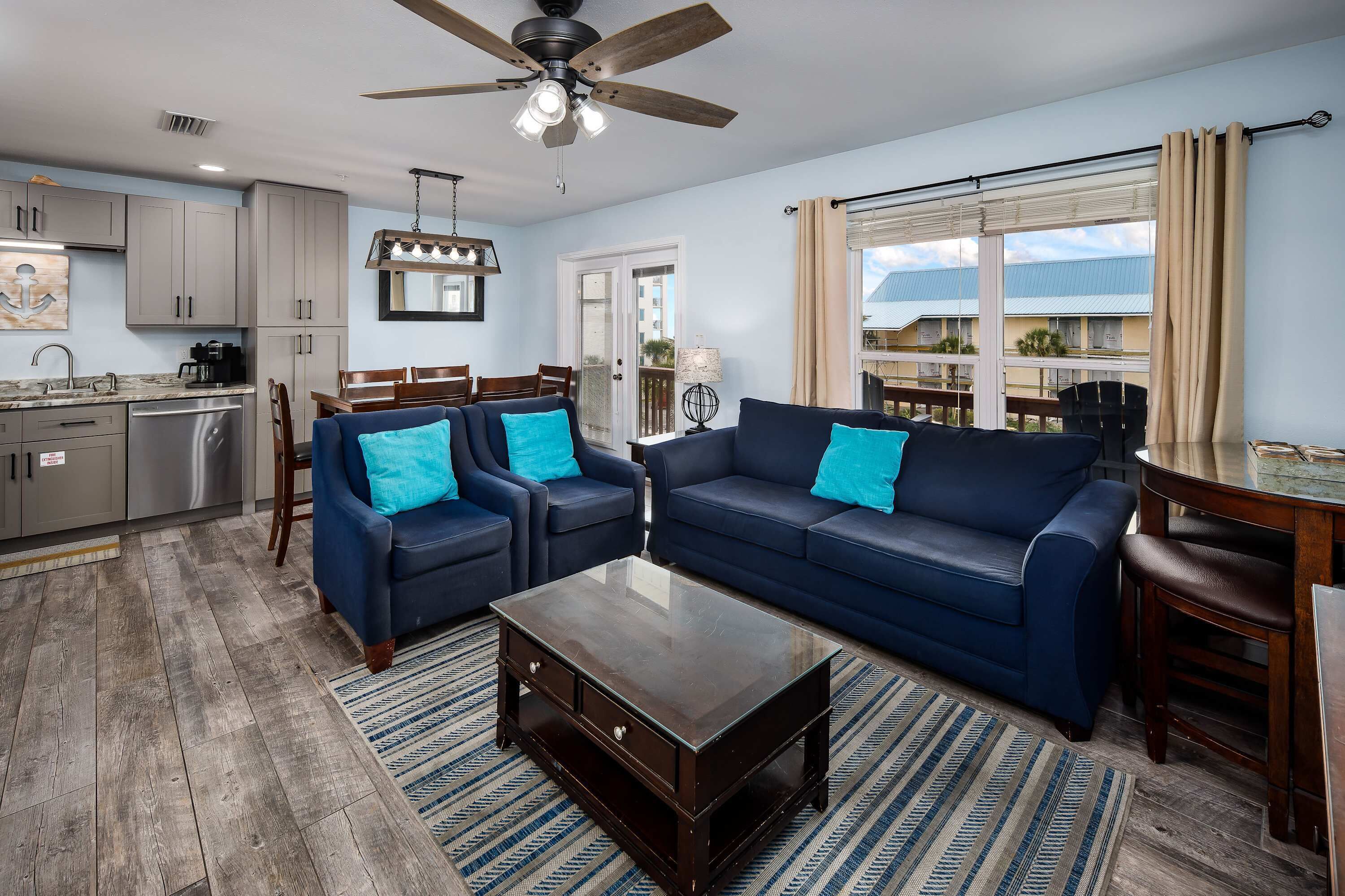 A modern living room with a dark wood coffee table, blue sofas with teal cushions, a dining area with a wooden table, and a large window with a view of buildings.