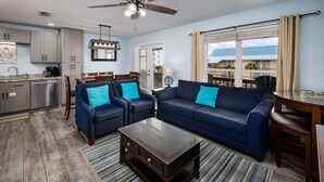 Ein modernes Wohnzimmer mit einem dunklen Holztisch, blauen Sofas mit türkisblauen Kissen, einem Essbereich mit Holztisch und einem großen Fenster mit Blick auf Gebäude.