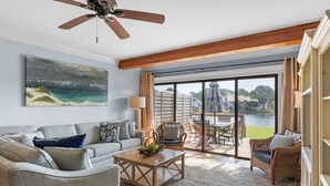 A spacious living room with a large window, a ceiling fan, a sofa, a coffee table, and a wicker chair. - Renovated Beach Villa w/GOLF CART, Lakeside, Beach, Pool/Hot Tub, Tram, Grill (Miramar Beach)
