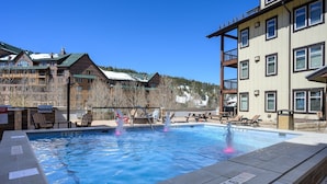 Uma piscina com uma fonte, rodeada por espreguiçadeiras e mesas de piquenique, com edifícios e uma montanha nevada ao fundo.