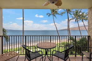 A balcony with a view of the ocean, palm trees, and a table with chairs.