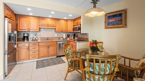 A kitchen with wooden cabinets, stainless steel appliances, a dining table with chairs, and a hanging pendant light. - Island Charm: Kihei Beach #305, Beachfront Condo Offers Spectacular Ocean Views! (Kihei, Maui)