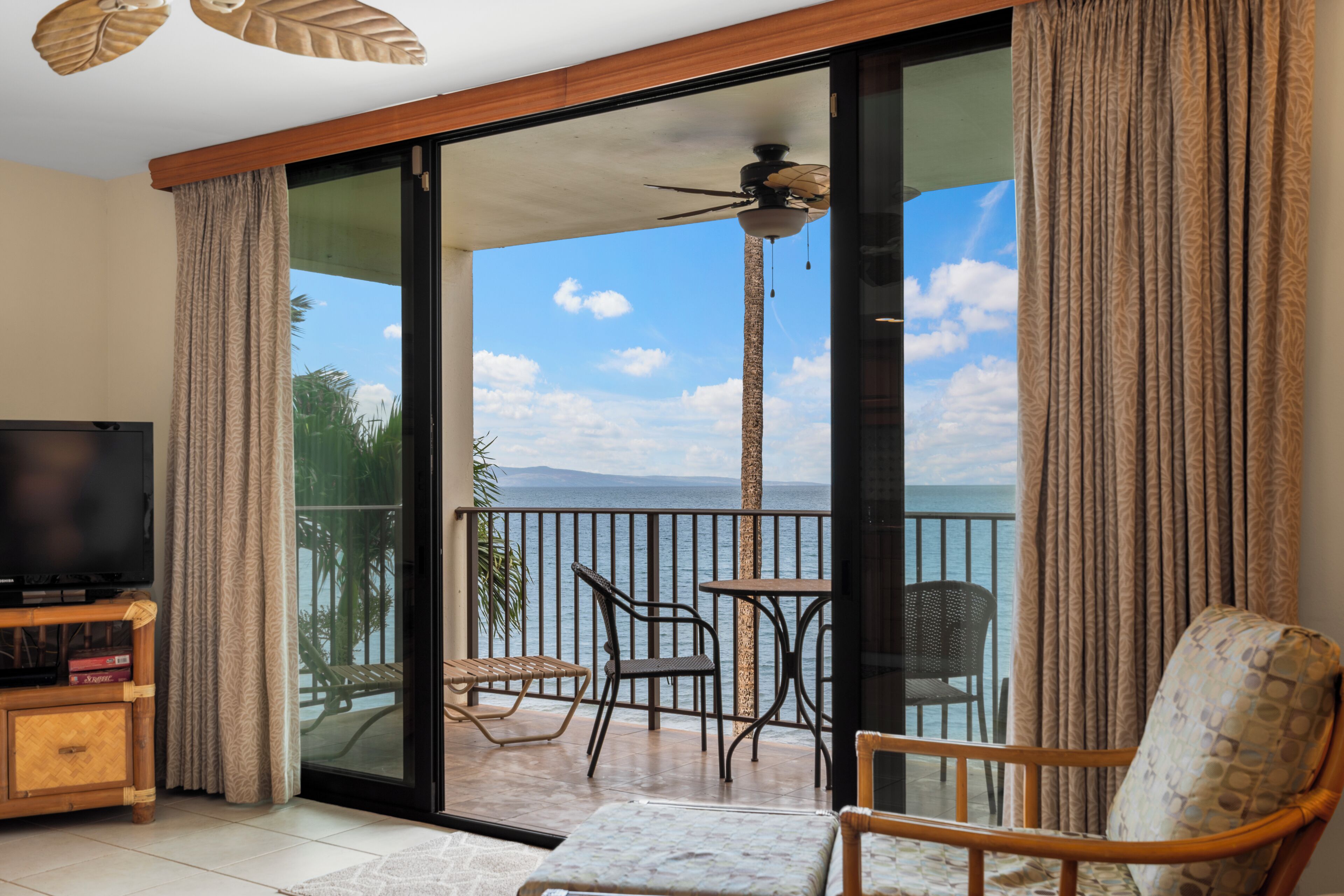 Een balkon met uitzicht op zee, een plafondventilator en een televisie op een houten kast.