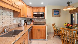 A kitchen with wooden cabinets, a stainless steel microwave, a sink, and a dining area with a table and chairs. - Island Charm: Kihei Beach #305, Beachfront Condo Offers Spectacular Ocean Views! (Kihei, Maui)