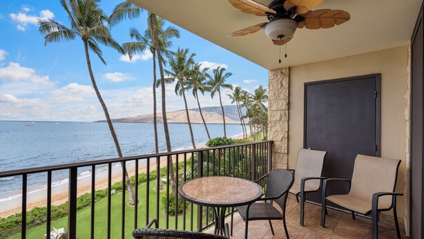 A balcony with a view of the ocean, palm trees, and a beach. There are two chairs and a table. - Island Charm: Kihei Beach #305, Beachfront Condo Offers Spectacular Ocean Views! (Kihei, Maui)