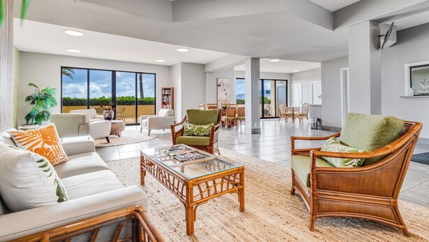 A spacious living area with a large glass coffee table, wicker furniture, and a view of the outdoors. - Island Charm: Kihei Beach #305, Beachfront Condo Offers Spectacular Ocean Views! (Kihei, Maui)