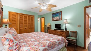 A bedroom with a bed, a ceiling fan, a television, a wooden wardrobe, and a chair. - Island Charm: Kihei Beach #305, Beachfront Condo Offers Spectacular Ocean Views! (Kihei, Maui)