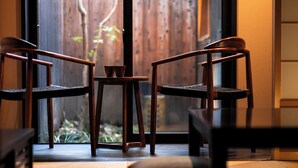 Une pièce avec deux chaises en bois et une petite table, une porte-fenêtre coulissante donnant sur une clôture en bois, et une vue sur la verdure à l’extérieur.