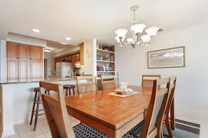 A dining area with a wooden table and chairs, a chandelier, and a kitchen in the background. - Ocean One 409 - Oceanfront 4th Floor Condo (Hilton Head Island)