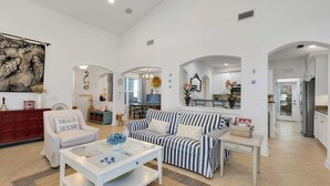 A spacious living room with a striped sofa, a white armchair, and a coffee table. There is a dining area with a table and chairs, and a kitchen visible through an archway. - Seagrove Beach Home + Carriage House + Bikes (Santa Rosa Beach)