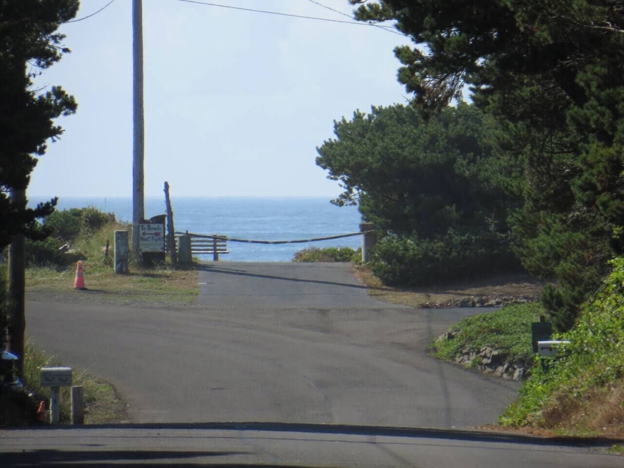 Une route côtière menant à l’entrée d’une plage, protégée par une barrière, et bordée de verdure, d’où l’on a une vue dégagée sur la mer.