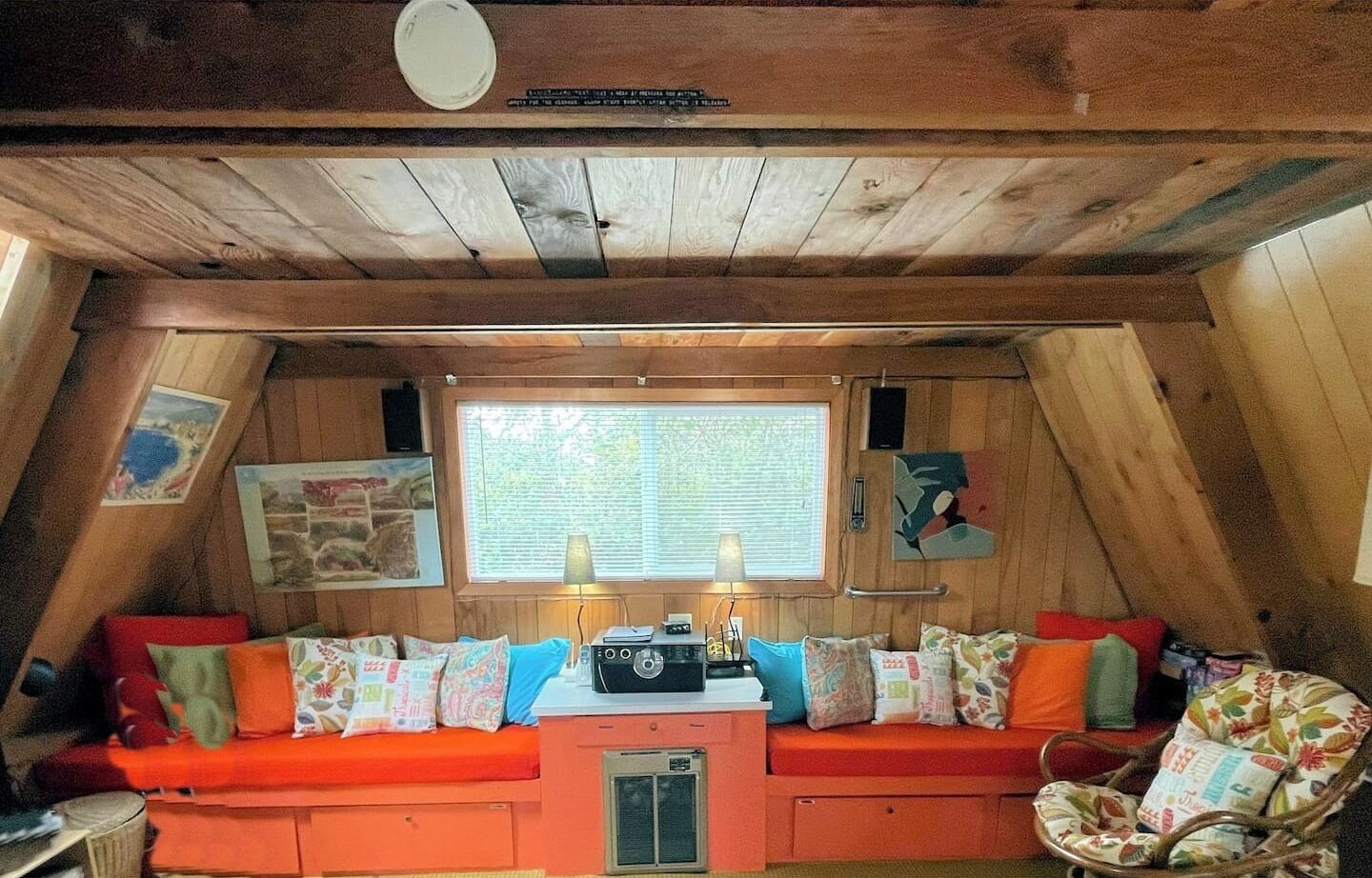 A cozy cabin interior with a wooden ceiling, a large window, a sofa with colorful cushions, and a small table with a radio.