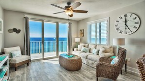 A spacious living room with a large window overlooking the sea, a ceiling fan, and a classic wall clock.