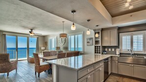A spacious kitchen with a large island, a dining area with wicker chairs, and a view of the ocean through the sliding glass doors.