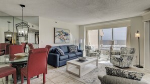 A spacious living room with a blue sofa, red and white patterned chairs, a coffee table, and a large window offering a view of the ocean.
