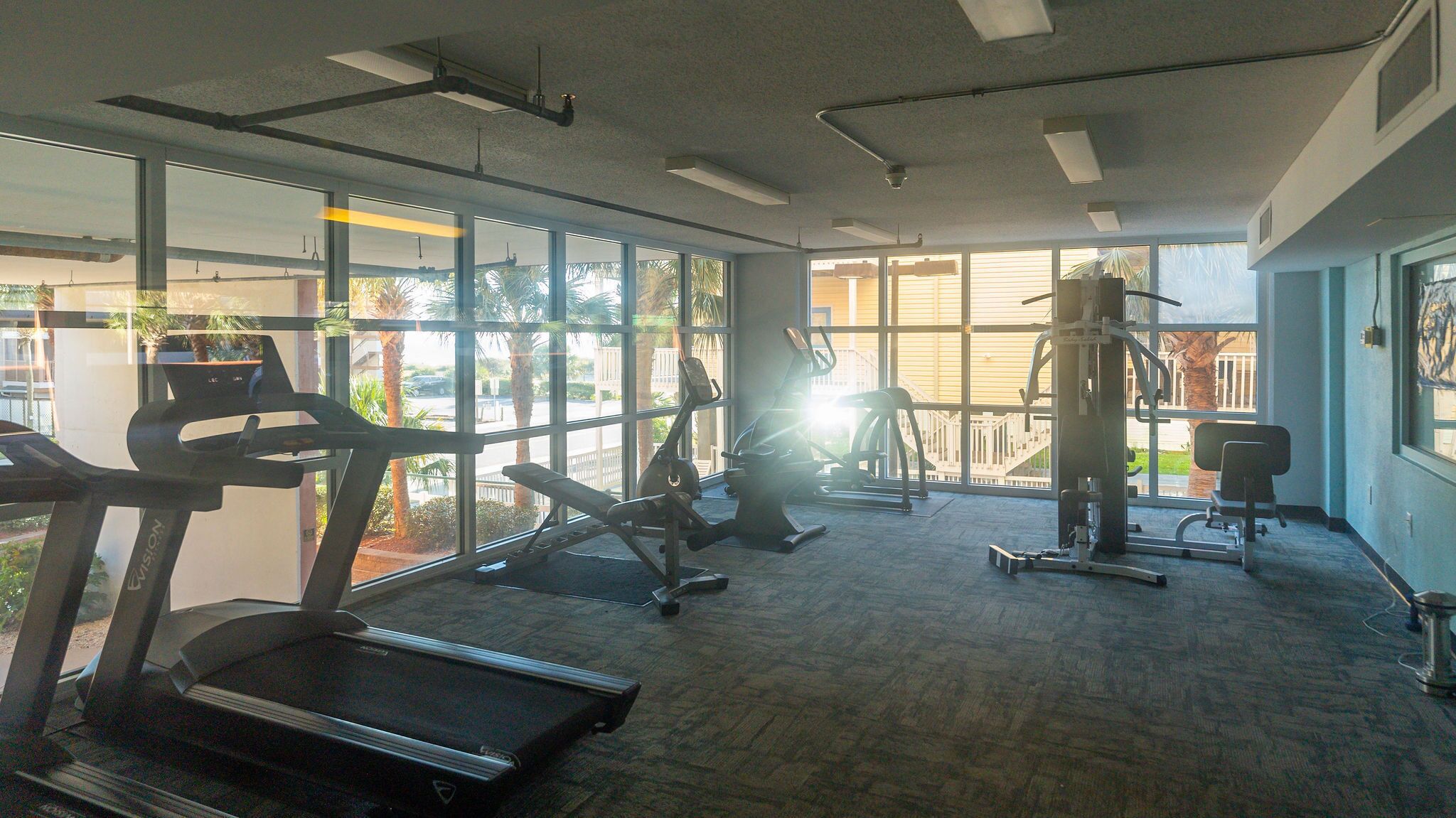 A gym with a treadmill, elliptical, and weight machine. Large windows provide natural light and a view of palm trees outside.