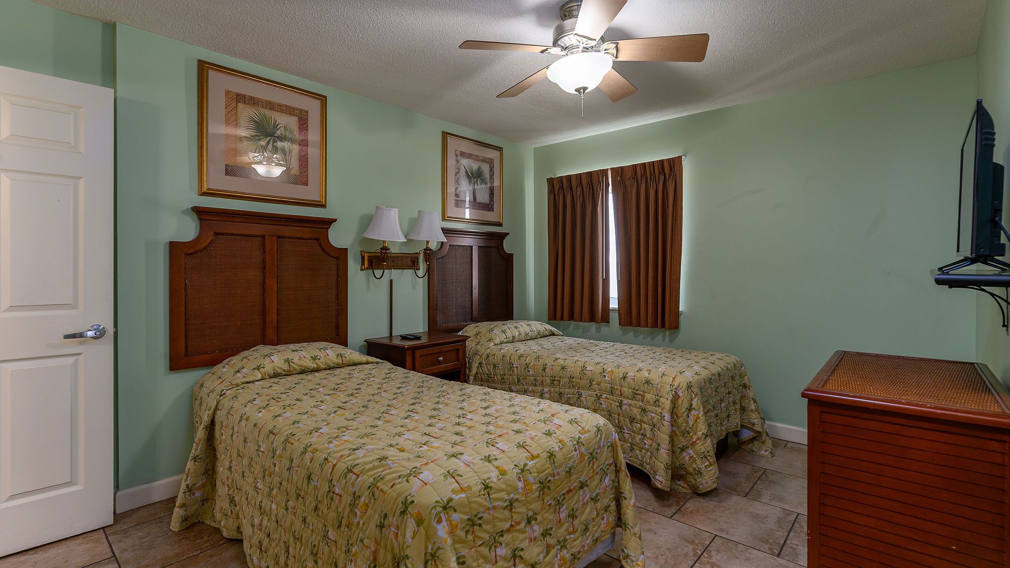Una habitación con dos camas, un ventilador de techo, una ventana con cortinas, un aparador de madera y cuadros enmarcados en la pared.