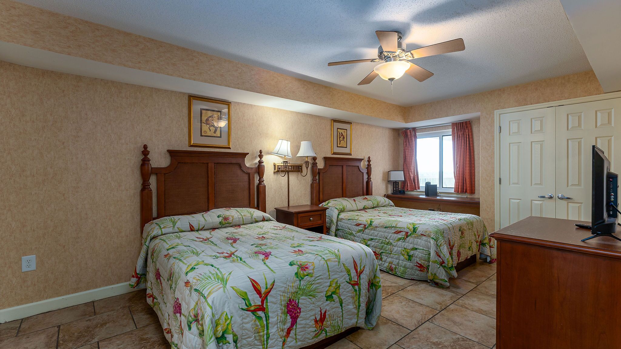 Quarto de hotel com duas camas, ventoinha no teto, escrivaninha com televisão e janela com cortinas.