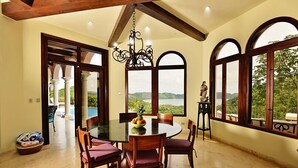A dining area with a round table, chairs, and a chandelier. There are large windows with arched tops, and a statue is visible on the right side.