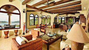 A spacious living area with wicker furniture, a glass coffee table, and large windows offering a view of the outdoors.