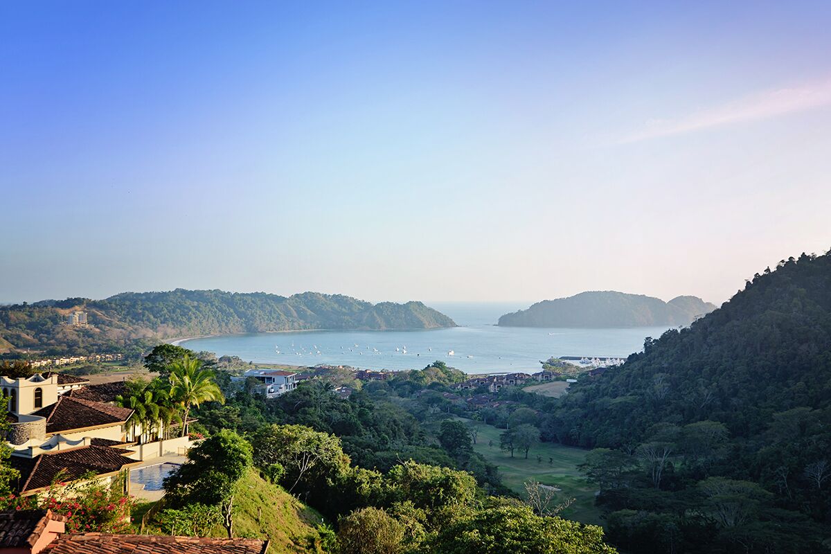 Paisaje costero con vista al mar, colinas y una zona residencial con casas y piscina.