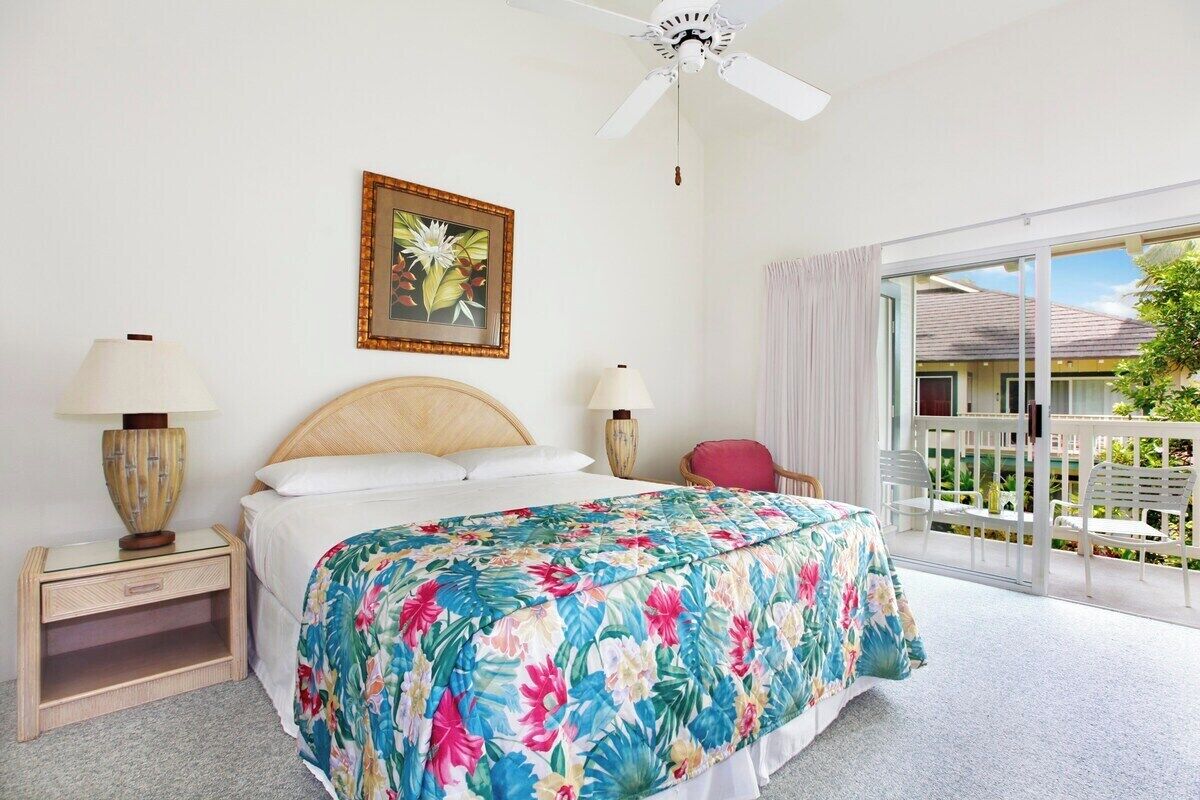 A bedroom with a bed, two bedside lamps, a ceiling fan, a window with curtains, and a balcony with chairs.