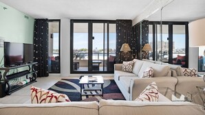 Una sala de estar moderna con sofá, mesa de centro y televisor. Cuenta con ventanas grandes con cortinas y vistas a la ciudad.