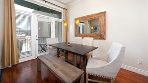 Un coin repas avec une table en bois, un banc et deux chaises rembourrées. Un grand miroir orne le mur, et une fenêtre est équipée de stores.