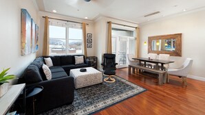 Un vaste salon comprenant un canapé noir, un pouf et une table à manger en bois avec des chaises. On y trouve une grande fenêtre avec des rideaux, un ventilateur de plafond et une œuvre d’art accrochée au mur.