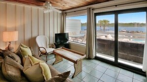 Una habitación con techo de madera, un sofá con cojines, una televisión, una mesa y vistas a un lago a través de amplias ventanas.