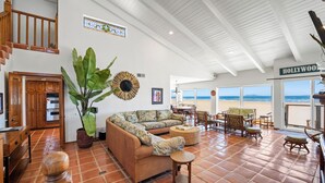 Un vaste espace de vie comprenant un grand canapé, une table à manger et des chaises. On y trouve également une plante en pot, un meuble en bois et un escalier menant à un niveau supérieur. Un panneau indiquant « HOLLYWOOD » est visible, et la pièce est dotée de grandes fenêtres offrant une vue sur la plage.