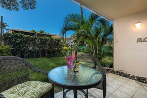 Una mesa redonda de vidrio con sillas de mimbre, una botella de vino y un florero con flores en un patio con vista a un jardín y un edificio al fondo.