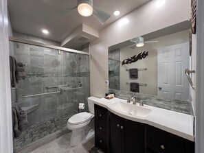 Un baño moderno con ducha de vidrio, inodoro blanco, tocador con espejo grande y ventilador de techo.