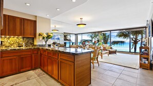 A spacious kitchen with wooden cabinets and a large island, a dining area with chairs, and a view of the ocean through large windows. - Timeless Island Charm with Stunning Ocean View (Lahaina)