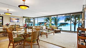 A spacious living area with a large glass window overlooking a beach, furnished with wicker chairs and a glass coffee table. - Timeless Island Charm with Stunning Ocean View (Lahaina)