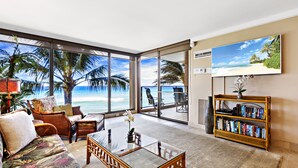 A spacious living room with a large glass door leading to a balcony with a view of the ocean. There is a comfortable sofa, a coffee table, and a bookshelf filled with books. - Timeless Island Charm with Stunning Ocean View (Lahaina)