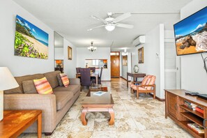 A living room with a sofa, coffee table, and TV. There is a ceiling fan, a dining area in the background, and a kitchen visible through an open door.