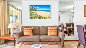 Una sala de estar con sofá, mesa de centro, mesa de comedor con sillas y un cuadro grande con temática playera en la pared.