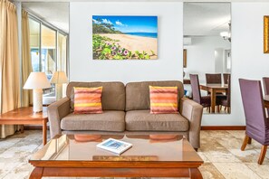Un soggiorno con divano, tavolino, tavolo da pranzo con sedie e un grande quadro a tema spiaggia.