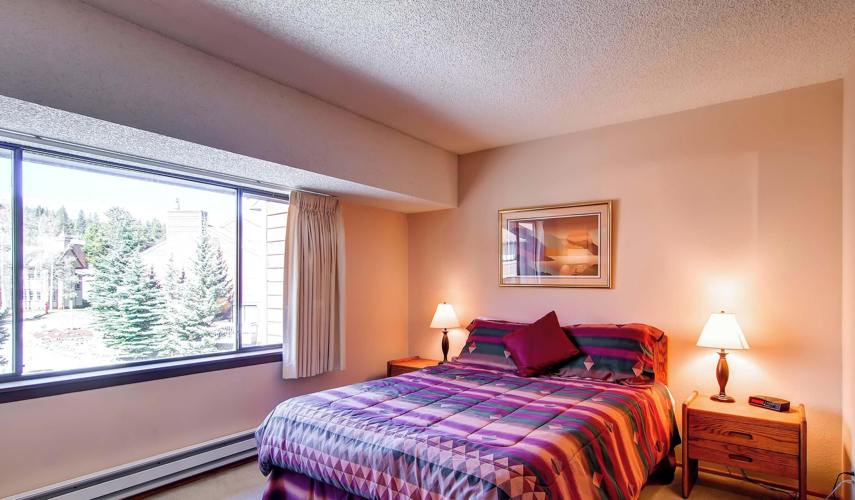 A bedroom with a large window, a bed with a colorful quilt, two bedside tables with lamps, and a framed picture on the wall.