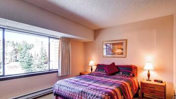 A bedroom with a large window, a bed with a colorful quilt, two bedside tables with lamps, and a framed picture on the wall.