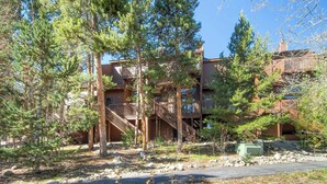 A multi-story building with a wooden staircase leading to a balcony, surrounded by trees and a rocky ground.