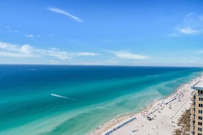 An aerial view of a beach with clear turquoise waters, a long stretch of sand, and a pier in the distance.