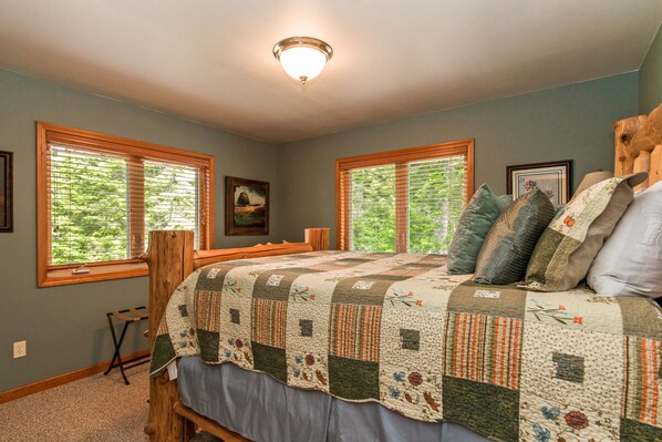 A bedroom with a large bed, two windows with blinds, a ceiling light, and a framed picture on the wall.