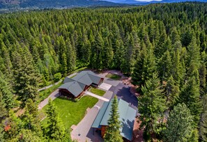 An aerial view of a cabin surrounded by dense forest, with a large deck area and a well-maintained lawn.