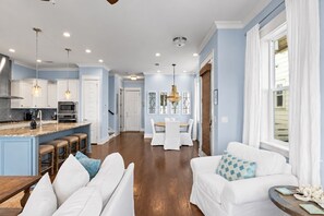 A modern kitchen with a central island, dining area with white chairs, and a living room with a white sofa.